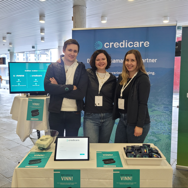 Tre representanter fra CrediCare står smilende foran en utstillingsstand med konkurranse og informasjonsmateriell på en messe.

